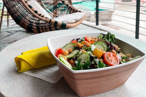 A square bowl of fresh salad with greens, cherry tomatoes, cucumber, and chickpeas sits on a round table next to a yellow napkin. In the background, there is a hanging chair and a poolside view.