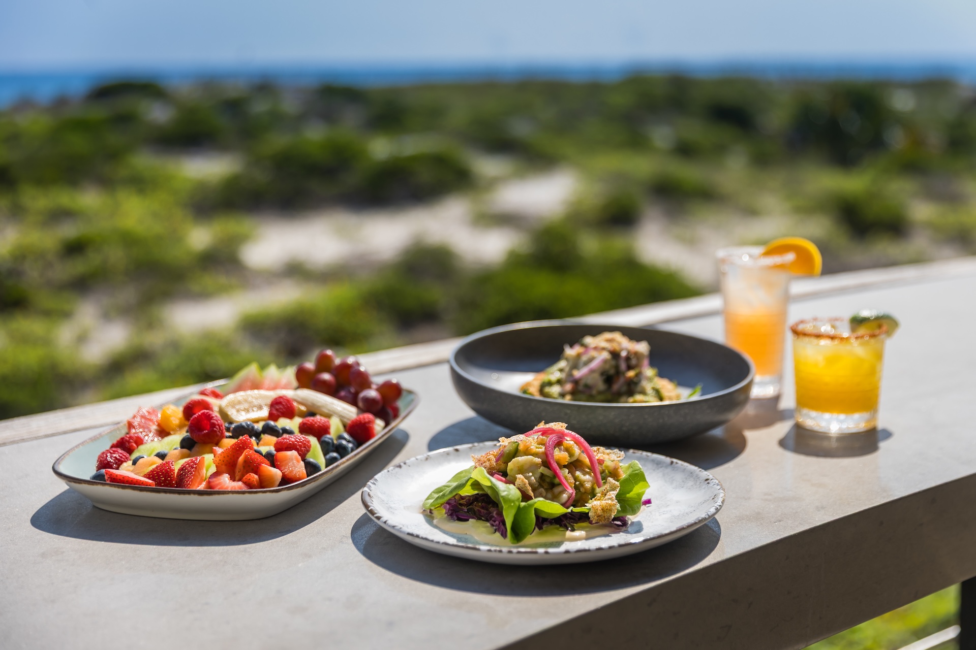 A table outdoors with plates of fresh fruit, salad, and tacos, alongside two colorful drinks. Greenery and a blue sky are visible in the background.