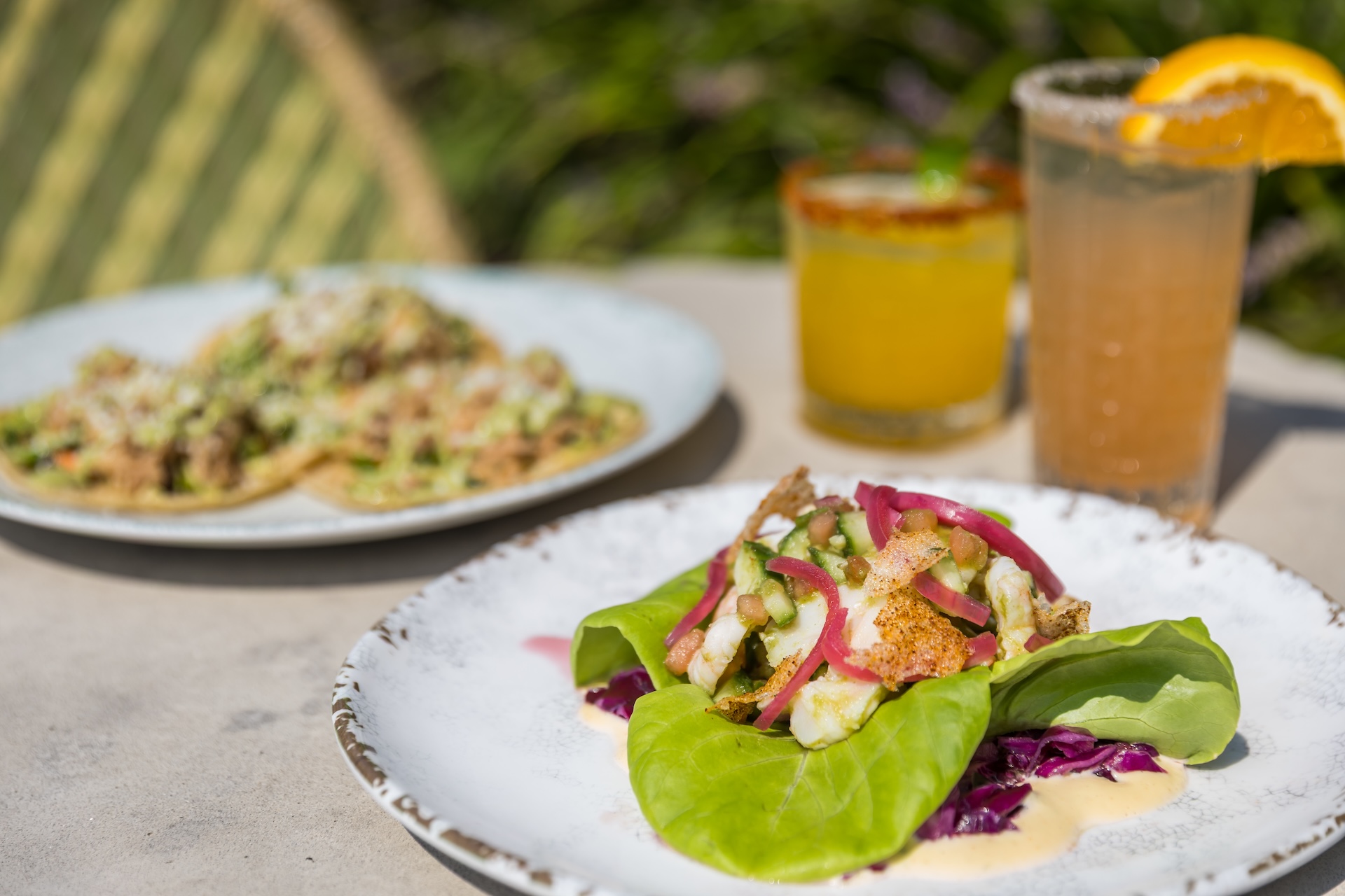 A close-up of a fresh lettuce wrap filled with grilled chicken, pickled onions, and sauce, on a white plate. In the background, tacos, a margarita, and a cocktail with an orange slice sit on an outdoor table.