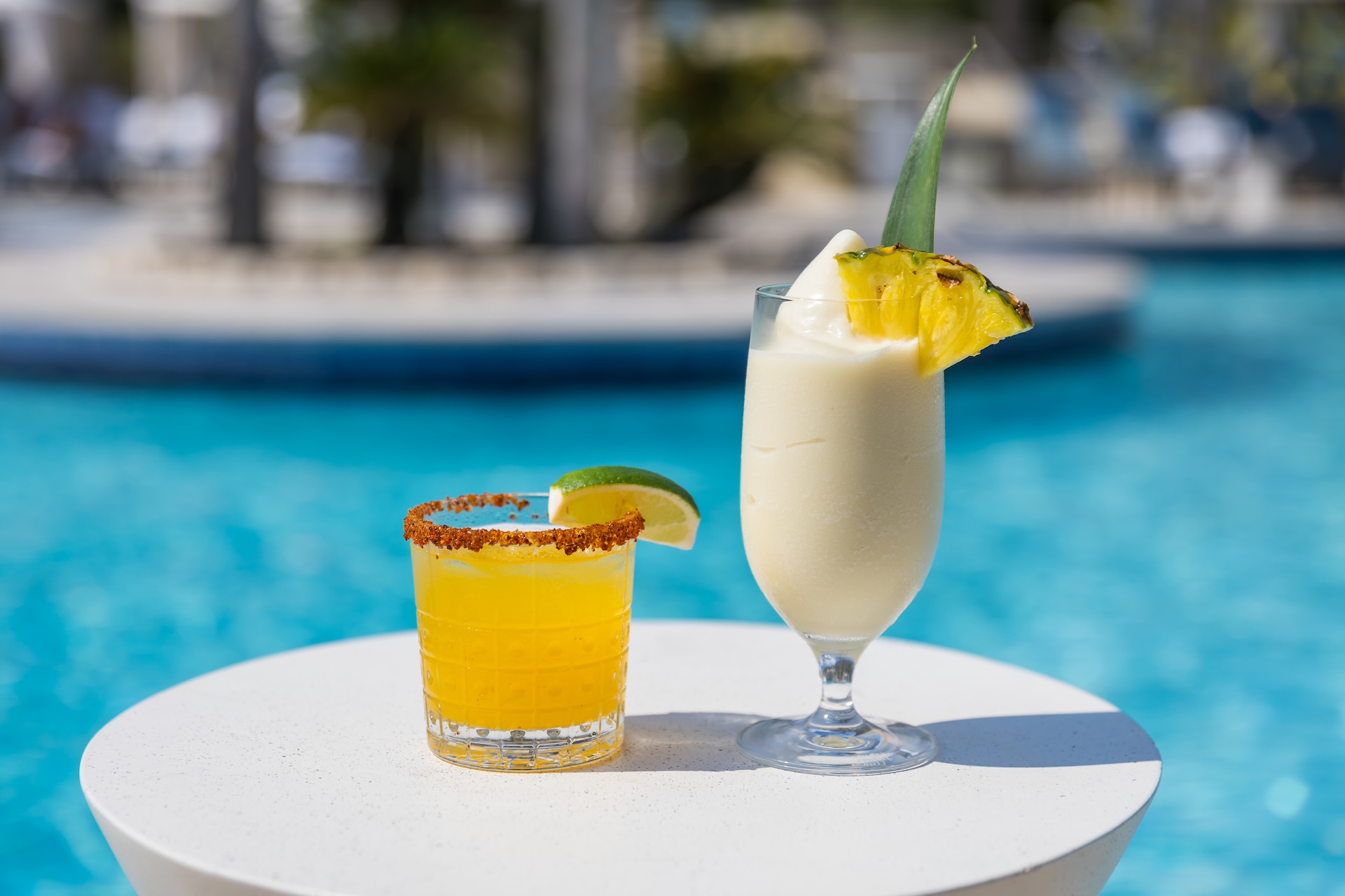 Two tropical cocktails on a round white table by a pool: one yellow drink with a lime wedge and chili rim, and one creamy drink with pineapple garnish and leaf, both with a blurred pool background.