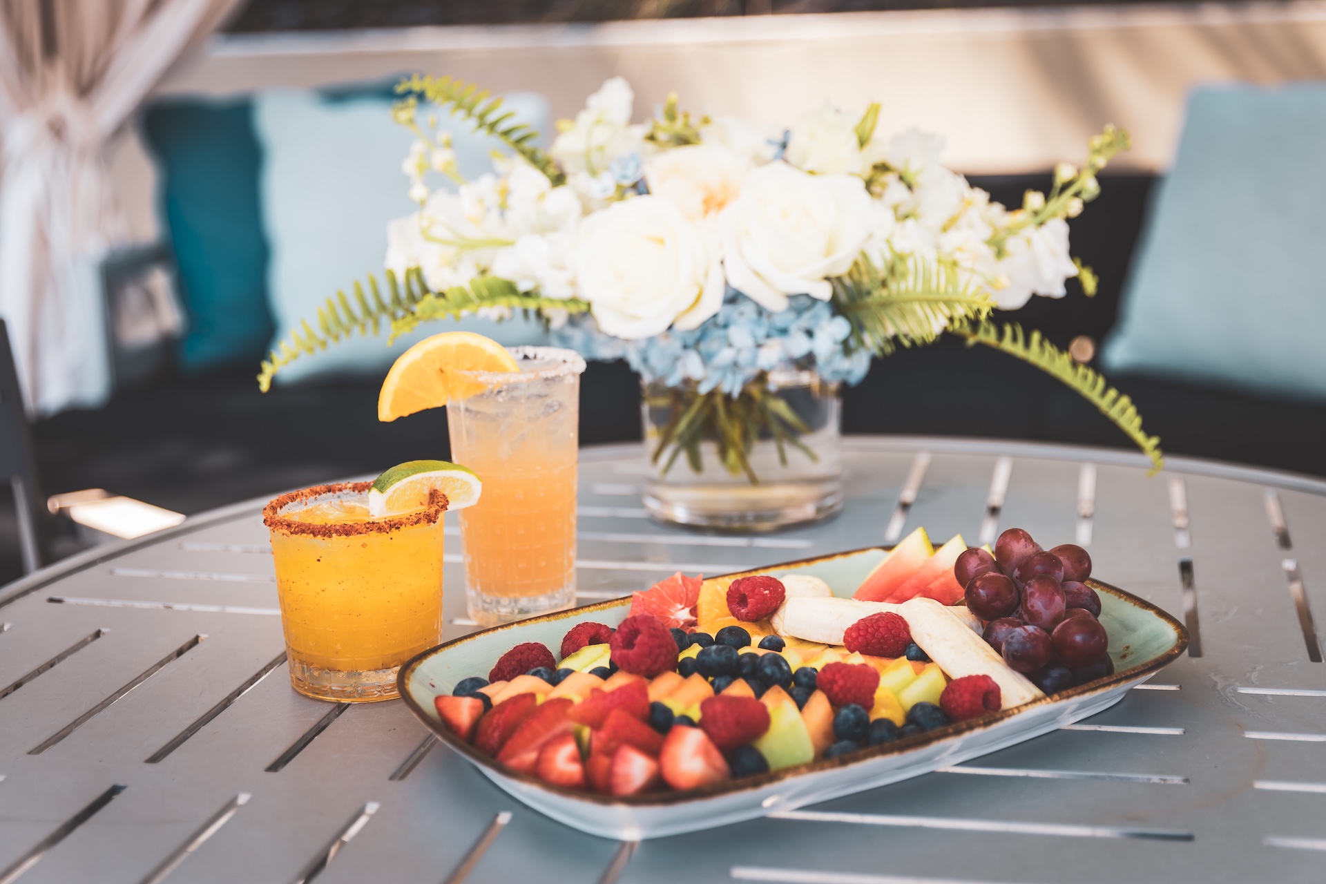 A plate of assorted fresh fruit sits on a round table beside two colorful drinks garnished with citrus slices; a vase of white and blue flowers is in the background.