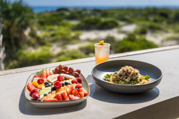 A plate of assorted fresh fruits and pancakes, a bowl of salad with a seafood topping, and a glass of orange drink on an outdoor ledge with greenery and the ocean in the background.