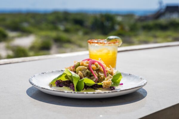 A plate of salad with greens, pickled onions, and croutons sits on a table outdoors, next to a yellow cocktail in a glass rimmed with chili powder and garnished with a lime wedge. Blurred greenery in the background.