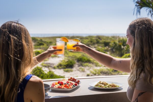 Two women clink glasses with orange drinks at an outdoor table overlooking greenery and a beach, with plates of fresh food in front of them under bright sunshine.