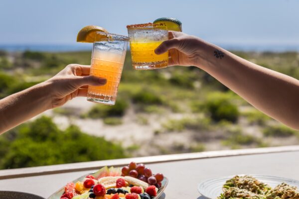Two people clink colorful cocktails garnished with fruit slices over a table with fresh fruit and salad, with a blurred outdoor landscape in the background.