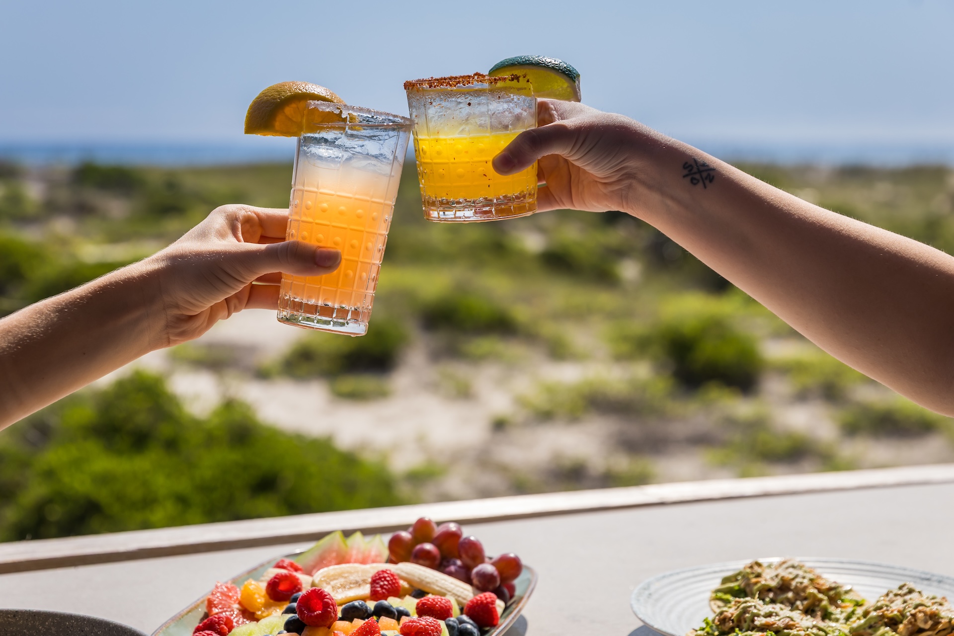 Two people clink colorful cocktails garnished with fruit slices over a table with fresh fruit and salad, with a blurred outdoor landscape in the background.
