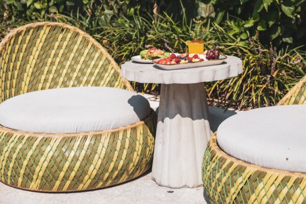 Two round wicker chairs with white cushions sit on either side of a small, round stone table outdoors. The table holds a tray with assorted fruits, cheese, and crackers. Lush greenery is in the background.