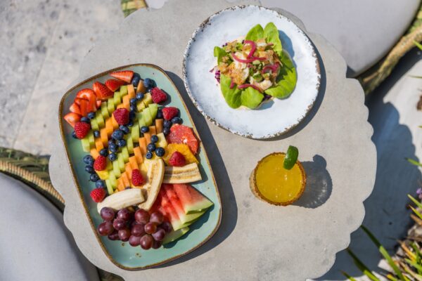 A tabletop with a colorful fruit platter, a plate with a salad on leafy greens, and a glass of orange juice garnished with a lime slice, set outdoors with wicker chairs partially visible.