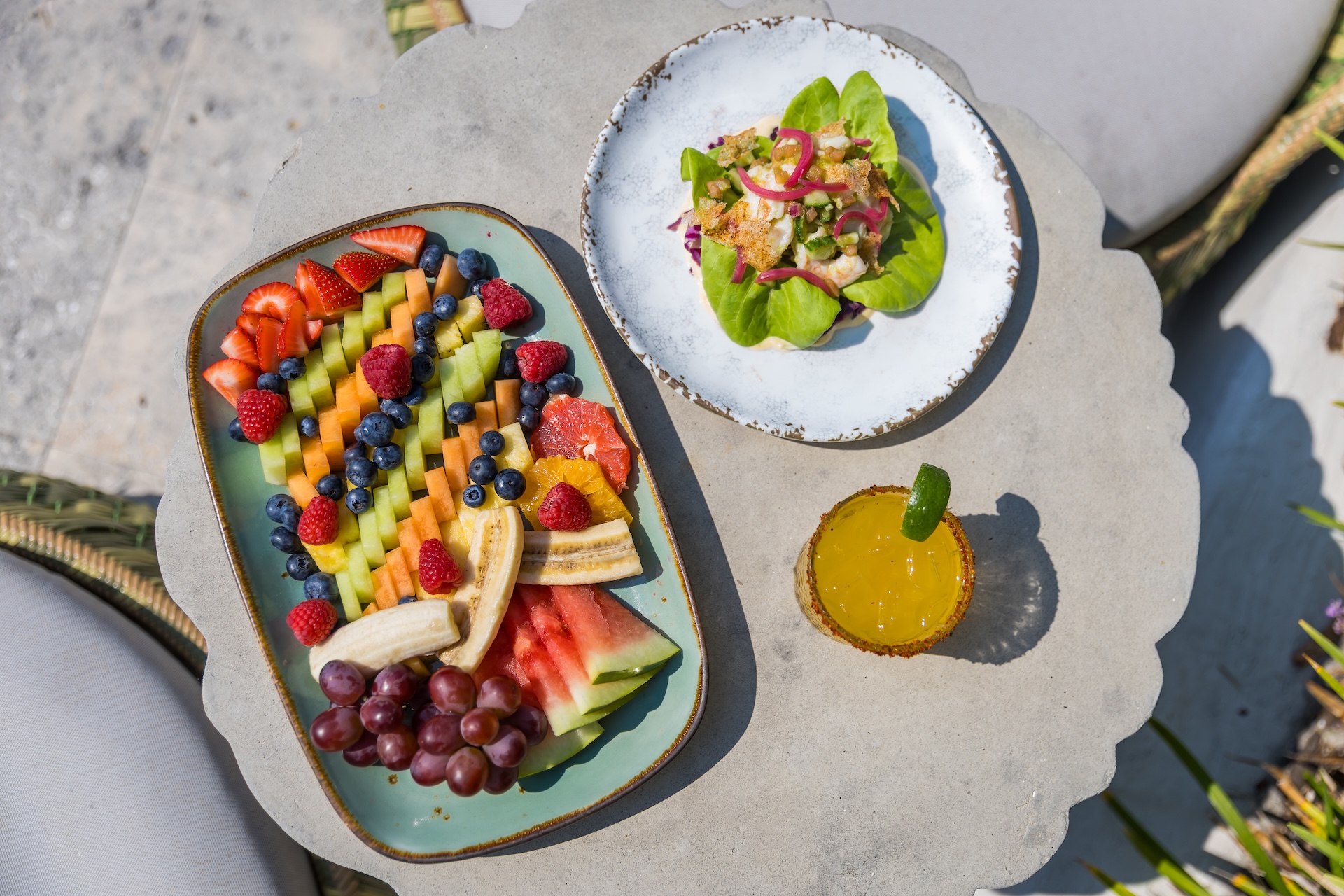 A tabletop with a colorful fruit platter, a plate with a salad on leafy greens, and a glass of orange juice garnished with a lime slice, set outdoors with wicker chairs partially visible.
