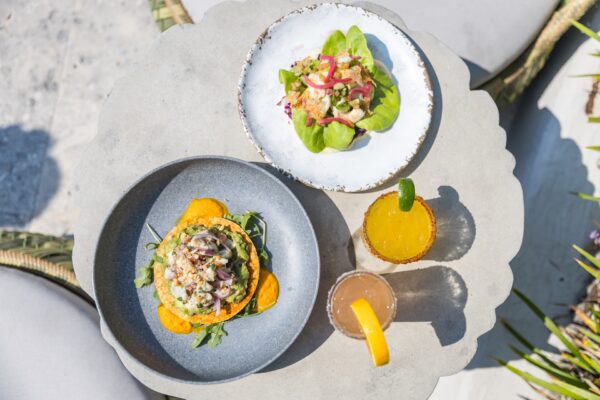 Top-down view of two plates with fresh salads, one served on a green leaf and the other with orange slices, accompanied by two colorful drinks on a light outdoor table.