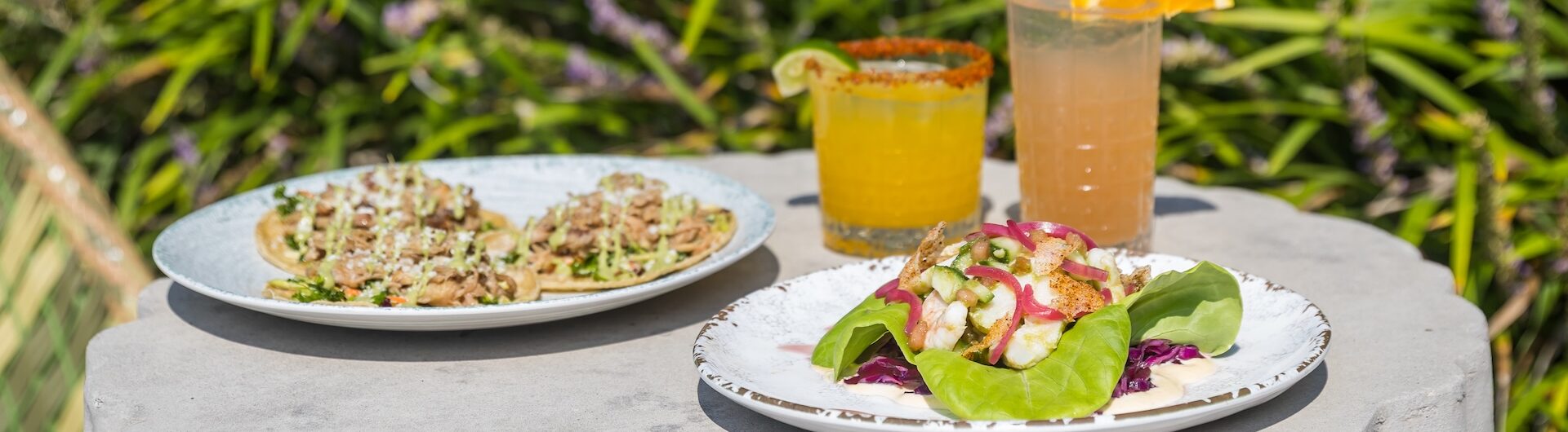 Two plates of food and two cocktails sit on a round stone table outside. One plate has tacos, the other has a leafy salad. The drinks are garnished with lime and orange. Green plants are visible in the background.