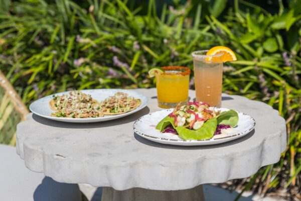 Two plates of food and two cocktails sit on a round stone table outside. One plate has tacos, the other has a leafy salad. The drinks are garnished with lime and orange. Green plants are visible in the background.