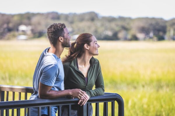 A man and a woman stand together outdoors, leaning on a railing and looking out at a field, smiling, on a sunny day.