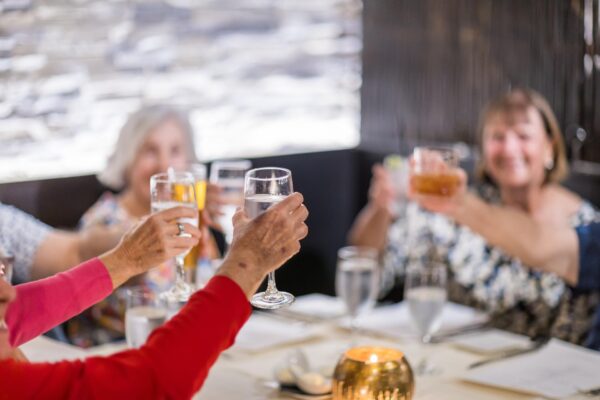 A group of people sitting at a restaurant table raise their glasses in a toast. The focus is on the raised glasses, with smiling faces blurred in the background.