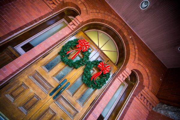 A wooden double door with arched windows is decorated with two green wreaths adorned with red and white bows. The building has red brick walls and an arched entryway.