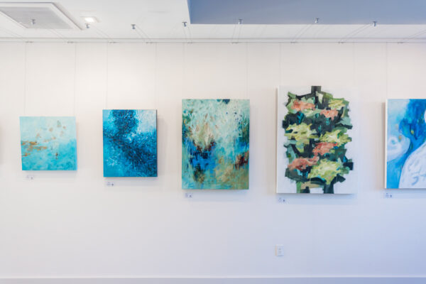 Five abstract paintings, mostly in blue and green tones, are displayed on a white wall in an art gallery with a minimal, modern setting.
