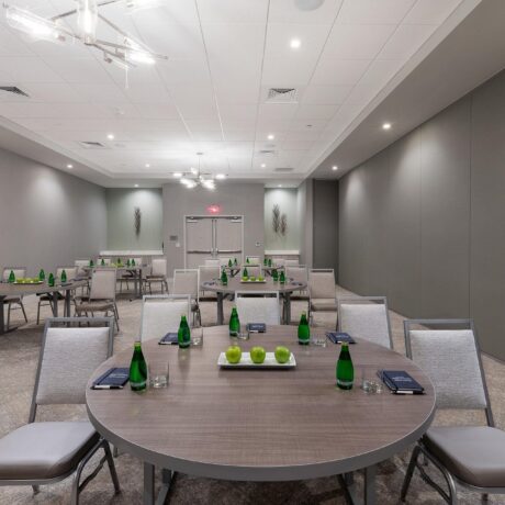 A modern conference room with round tables, green apples, bottled water, notepads, and chairs set up, under bright ceiling lights with neutral gray walls.