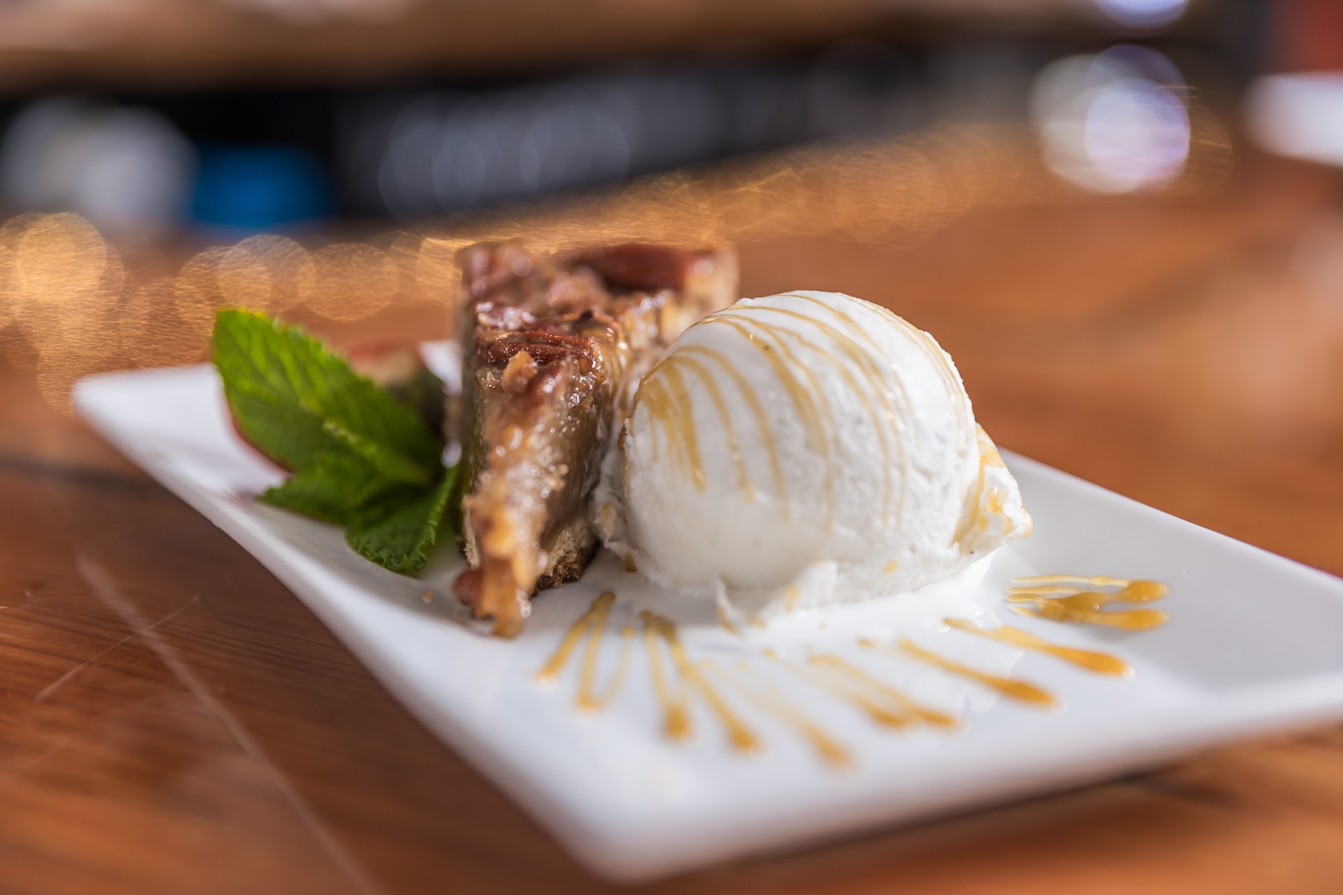 A slice of pecan pie garnished with mint next to a scoop of vanilla ice cream drizzled with caramel sauce on a rectangular plate.