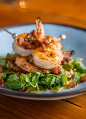 Two grilled shrimp on skewers served over a bed of arugula and bacon bits in a blue bowl.