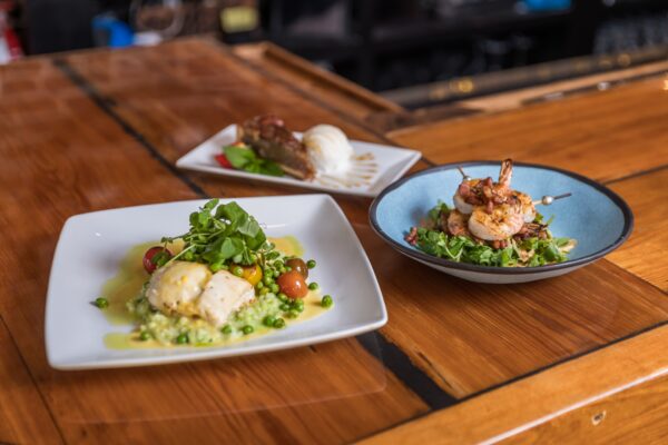 Three plates of food on a wooden table: a fish fillet with green risotto and tomatoes, a bowl of shrimp with greens, and a slice of dessert with a scoop of ice cream.