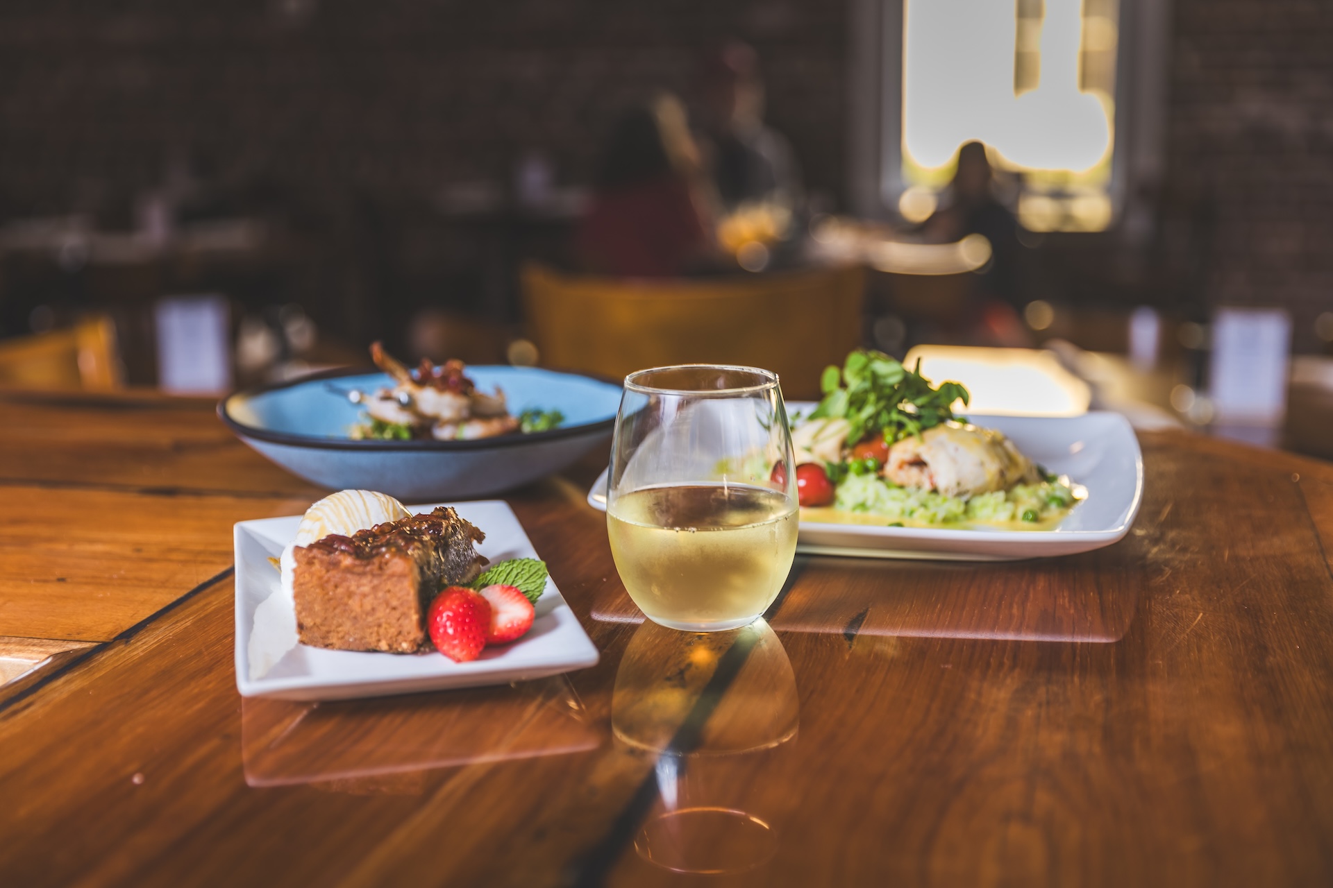 A slice of cake with ice cream and strawberries, a glass of white wine, and two plates of prepared food on a wooden table.