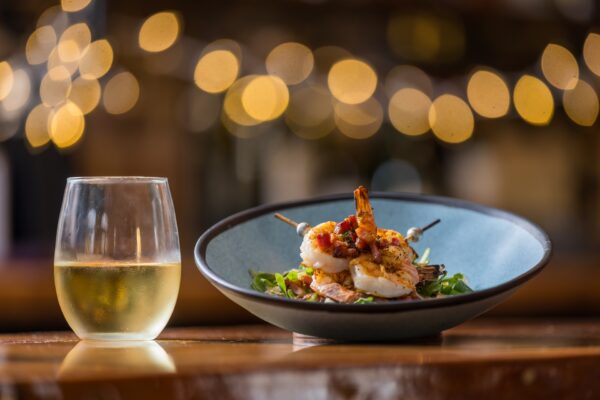 A bowl of shrimp skewers over greens next to a glass of white wine.