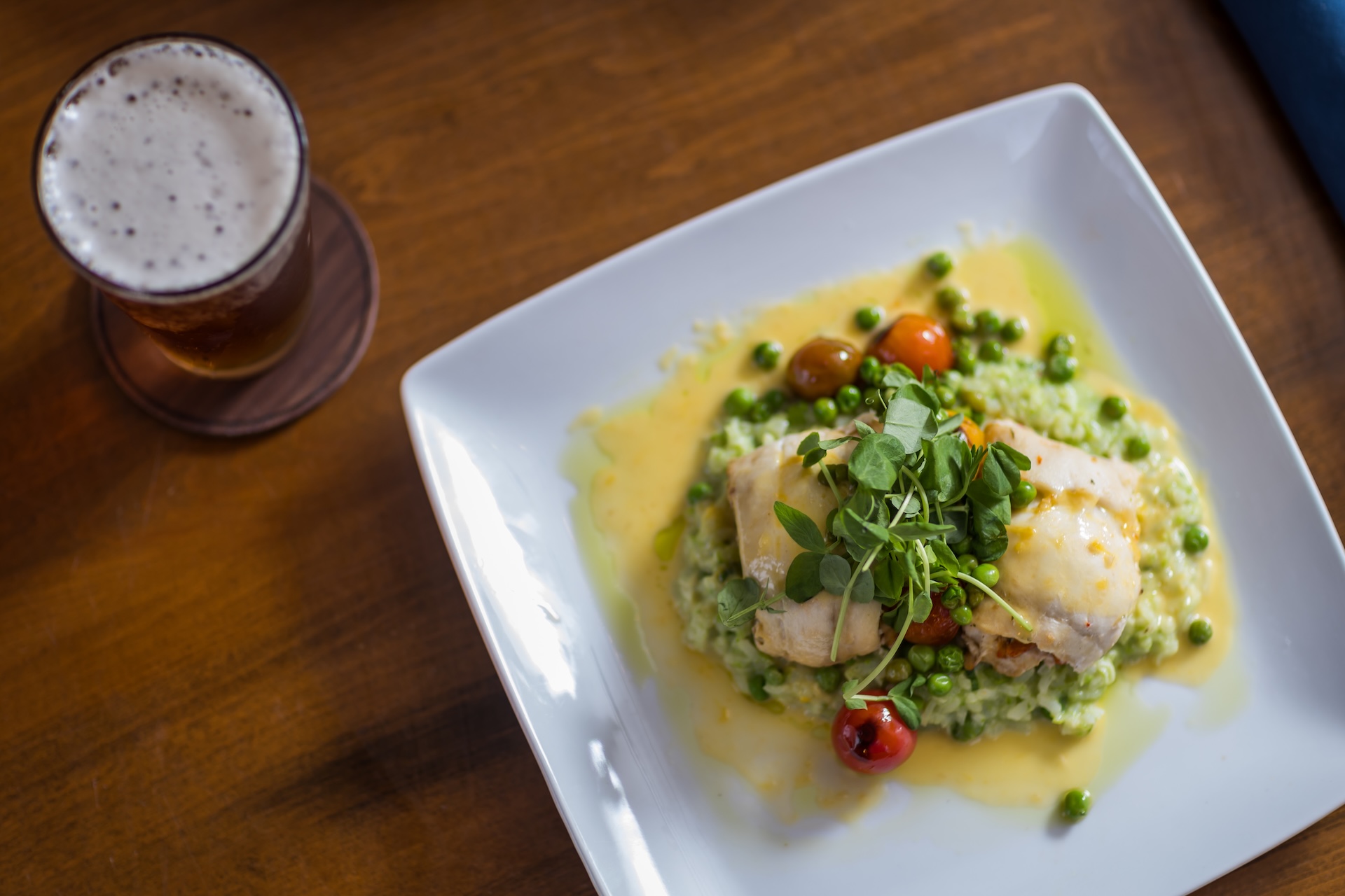 A white plate with chicken, green risotto, peas, and cherry tomatoes, garnished with greens, next to a glass of beer.