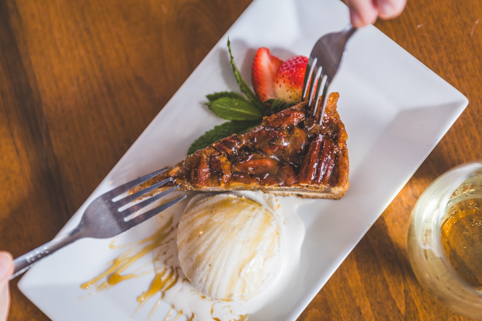 A slice of pecan pie with a scoop of vanilla ice cream, garnished with strawberries and mint, being eaten with a fork.