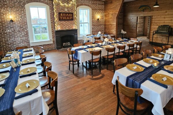 A cozy dining room with brick walls and wood floors, set with long tables covered in white cloths, blue runners, and gold plates. String lights and a sign reading CRAB TRAP hang above a fireplace.