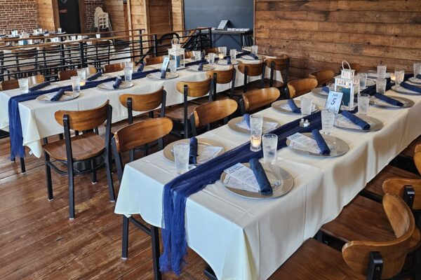 Two long banquet tables set for an event with white tablecloths, navy blue napkins, plates, menus, clear cups, and table numbers. Wooden chairs surround the tables in a rustic, wood-paneled room.