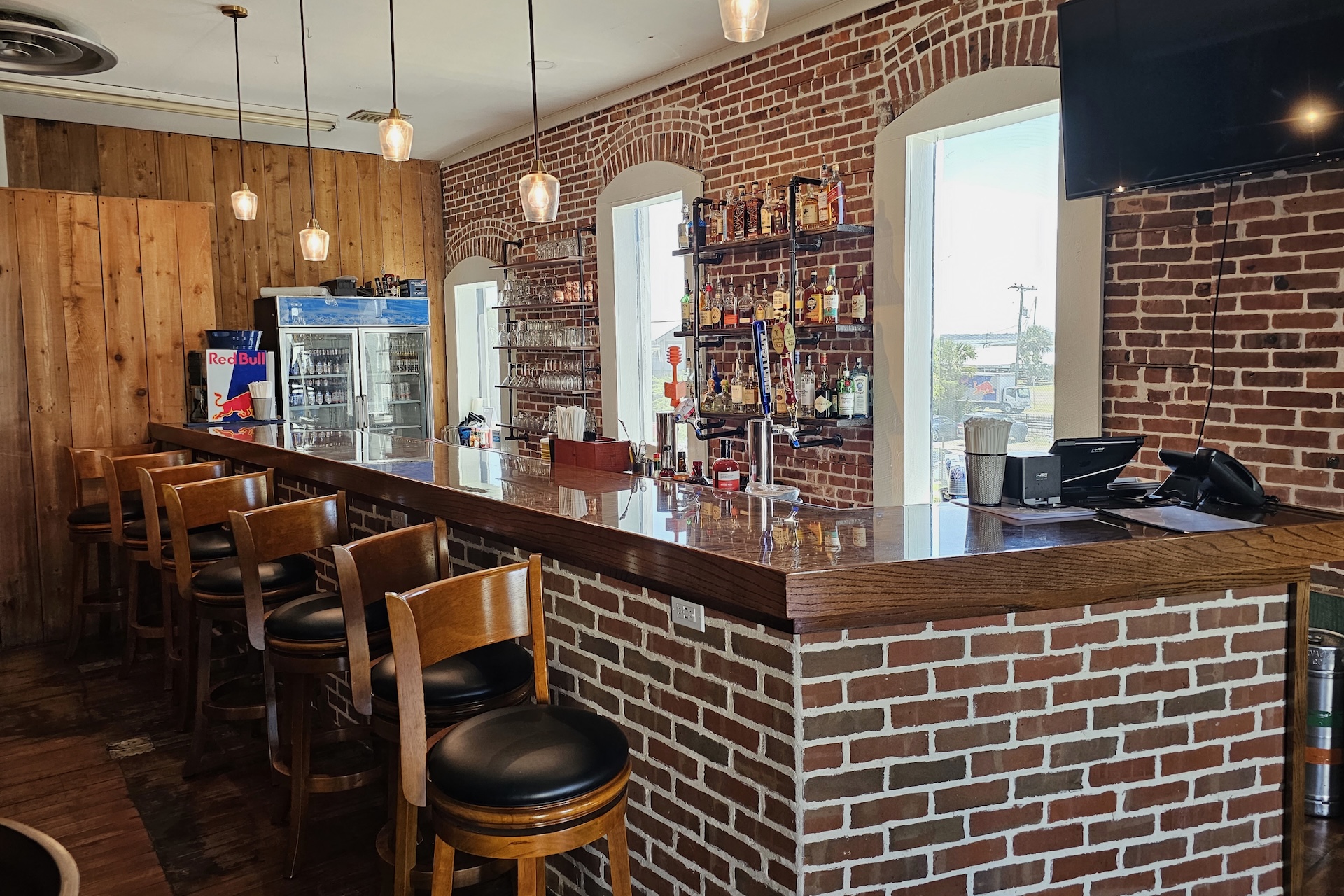 A cozy bar with brick walls, a wooden countertop, high wooden chairs, shelves stocked with liquor bottles, a refrigerator with drinks, large windows, and hanging pendant lights.