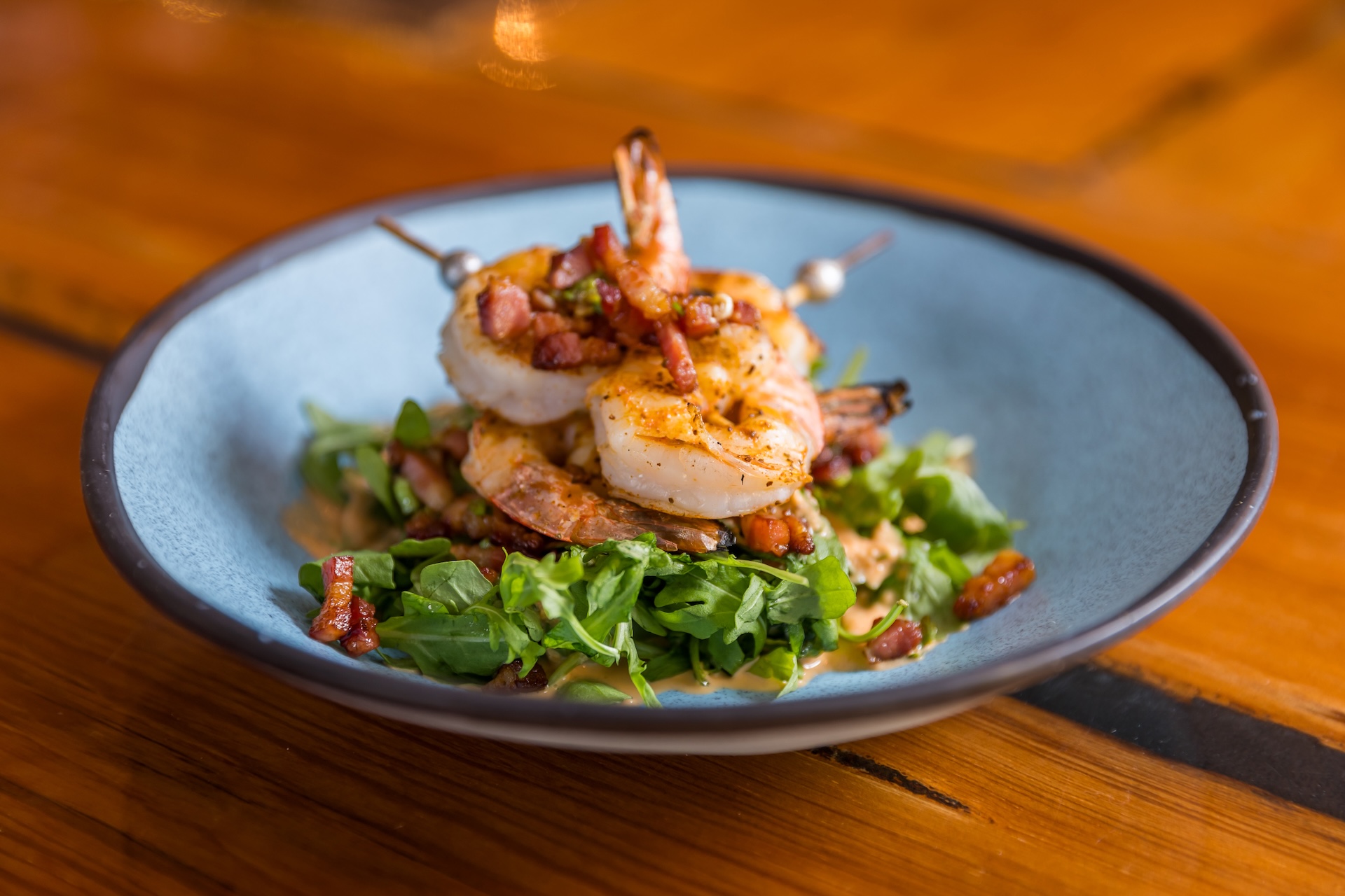 Two grilled shrimp on skewers served over a bed of arugula and bacon bits in a blue bowl.