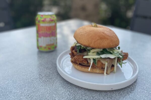 A fried chicken sandwich topped with slaw sits on a white plate on a gray table. A colorful can of sparkling beverage is in the background. The scene is outdoors with blurred greenery.