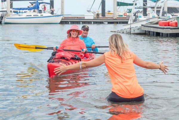 Two smiling people in a red kayak paddle toward a woman standing in shallow water with open arms, at a marina with several boats docked in the background.