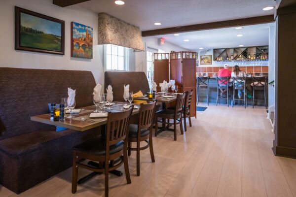 A cozy restaurant interior with brown booth seating, neatly set tables with glasses and napkins, wall art, and a bar area in the background with bottles and two people sitting on stools.