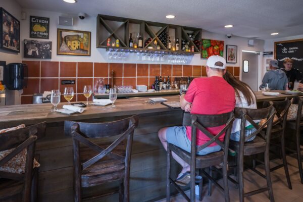 Two people sit at a wooden bar in a cozy restaurant, facing shelves with bottles and glasses. The walls are decorated with colorful art and framed pictures. Wine glasses and napkins are set on the counter.