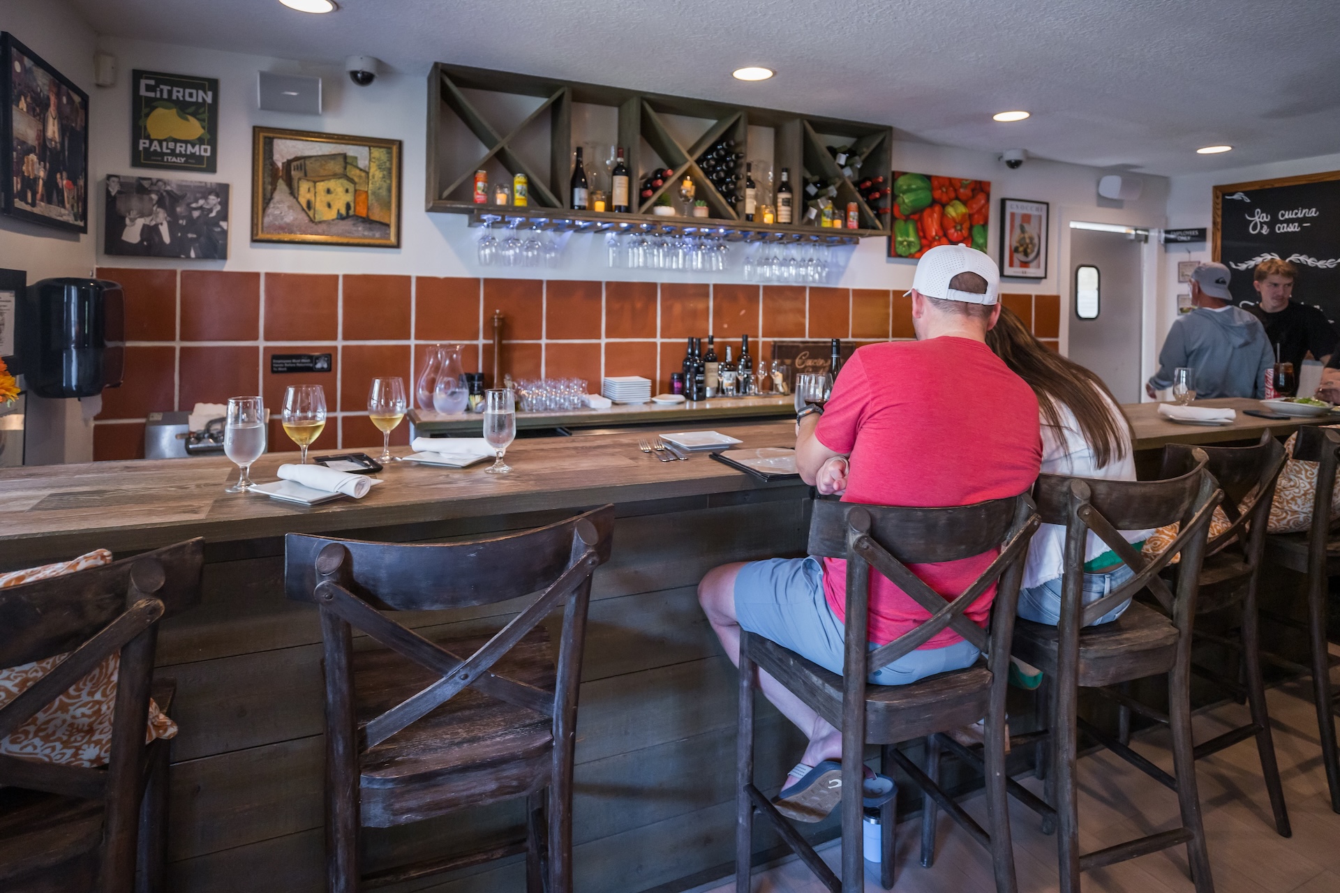 Two people sit at a wooden bar in a cozy restaurant, facing shelves with bottles and glasses. The walls are decorated with colorful art and framed pictures. Wine glasses and napkins are set on the counter.