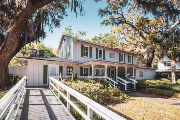 A two-story white wooden building with green shutters sits among large trees draped with Spanish moss. A ramp and stairs lead to the entrance, surrounded by shrubs and greenery on a sunny day.