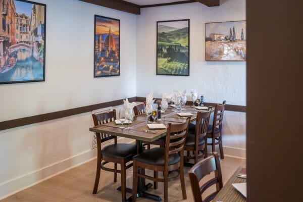 A cozy restaurant dining area with neatly set tables, wooden chairs, white napkins, and condiments. Framed pictures of scenic landscapes and cityscapes decorate the light-colored walls.