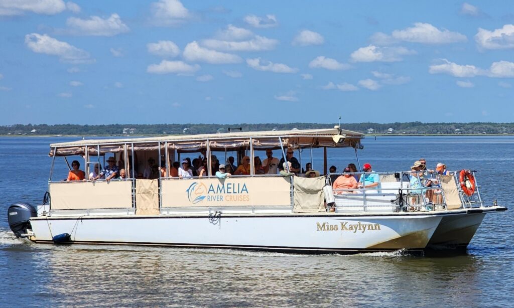A white sightseeing boat named Miss Kaylynn from Amelia River Cruises sails on calm water under a blue sky with scattered clouds, carrying several passengers seated under a canopy.