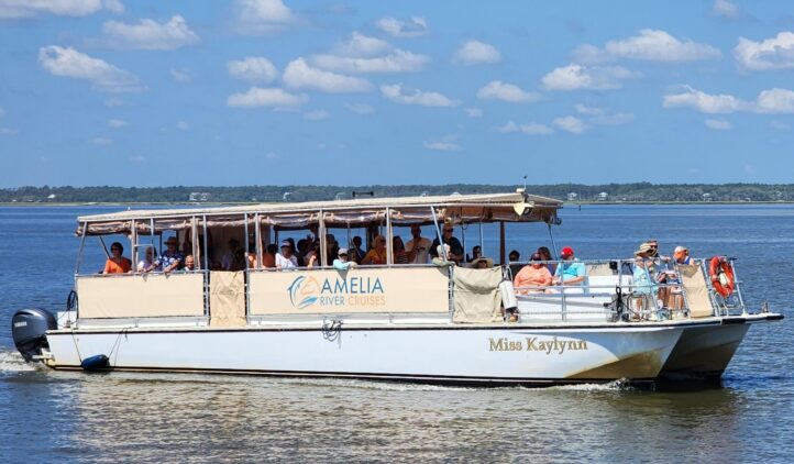 A white sightseeing boat named Miss Kaylynn from Amelia River Cruises sails on calm water under a blue sky with scattered clouds, carrying several passengers seated under a canopy.