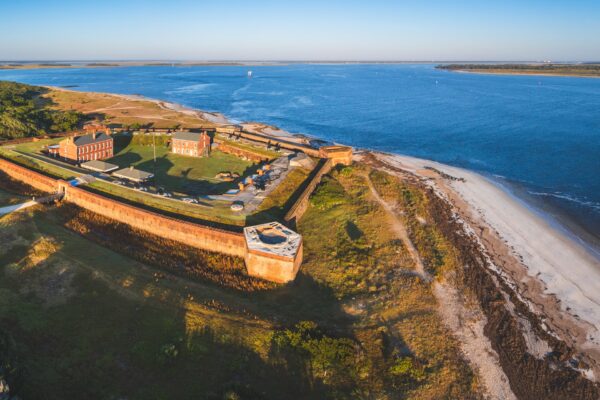 Aerial view of a historic brick fort on the coast, surrounded by grassy land and sandy beach, with blue ocean and distant shorelines in the background under a clear sky.
