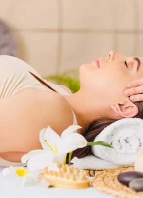 A woman lies on her back with eyes closed, receiving a head massage at a spa, surrounded by wellness items like flowers, herbal compresses, massage stones, and oil bottles.