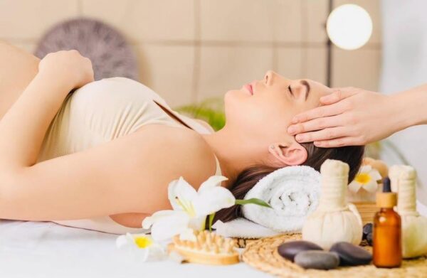 A woman lies on her back with eyes closed, receiving a head massage at a spa, surrounded by wellness items like flowers, herbal compresses, massage stones, and oil bottles.