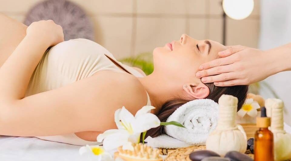 A woman lies on her back with eyes closed, receiving a head massage at a spa, surrounded by wellness items like flowers, herbal compresses, massage stones, and oil bottles.