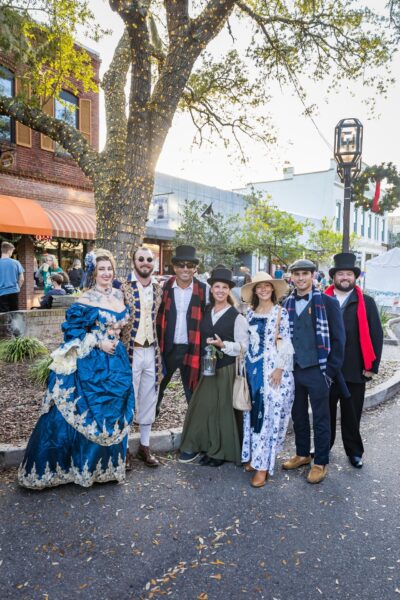 A group of seven people dressed in colorful Victorian-era costumes pose and smile outside on a city street, with shops, trees, and other people visible in the background.