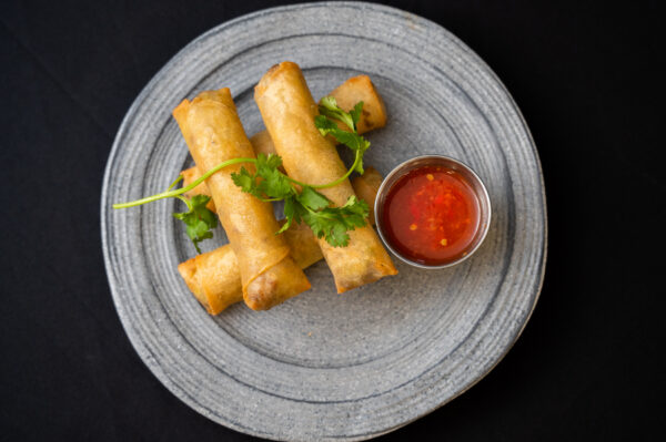 Three golden spring rolls garnished with fresh cilantro are arranged on a gray plate beside a small metal cup of red dipping sauce, set against a dark background.