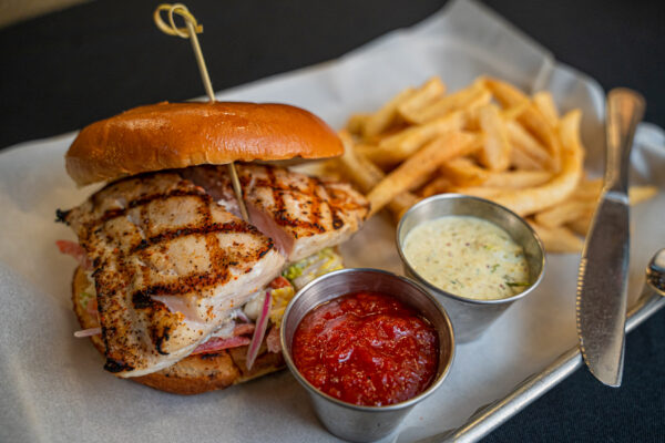 A grilled fish sandwich with lettuce and tomato on a brioche bun, served with a side of French fries, ketchup, and a creamy dipping sauce on a metal tray. A knife rests on the tray next to the food.
