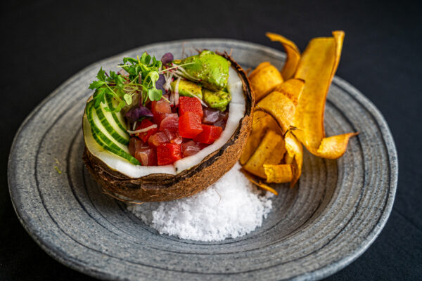 A halved coconut shell filled with diced raw fish, avocado, cucumber, and microgreens sits on a gray plate with a bed of salt, accompanied by plantain chips on the side.