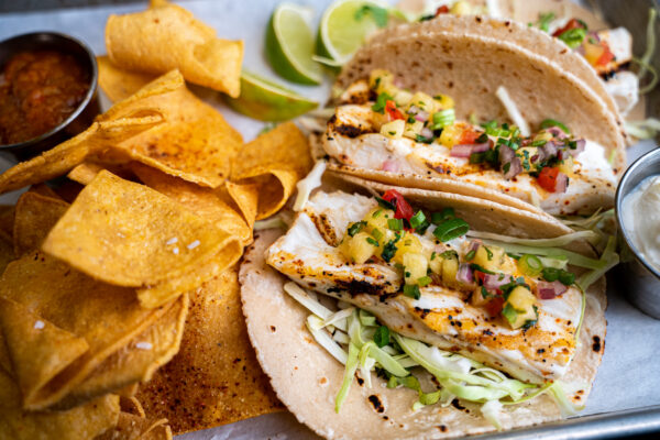 Two grilled fish tacos with cabbage, pineapple salsa, and scallions on tortillas, served with tortilla chips, lime wedges, salsa, and a side of sauce on a metal tray.